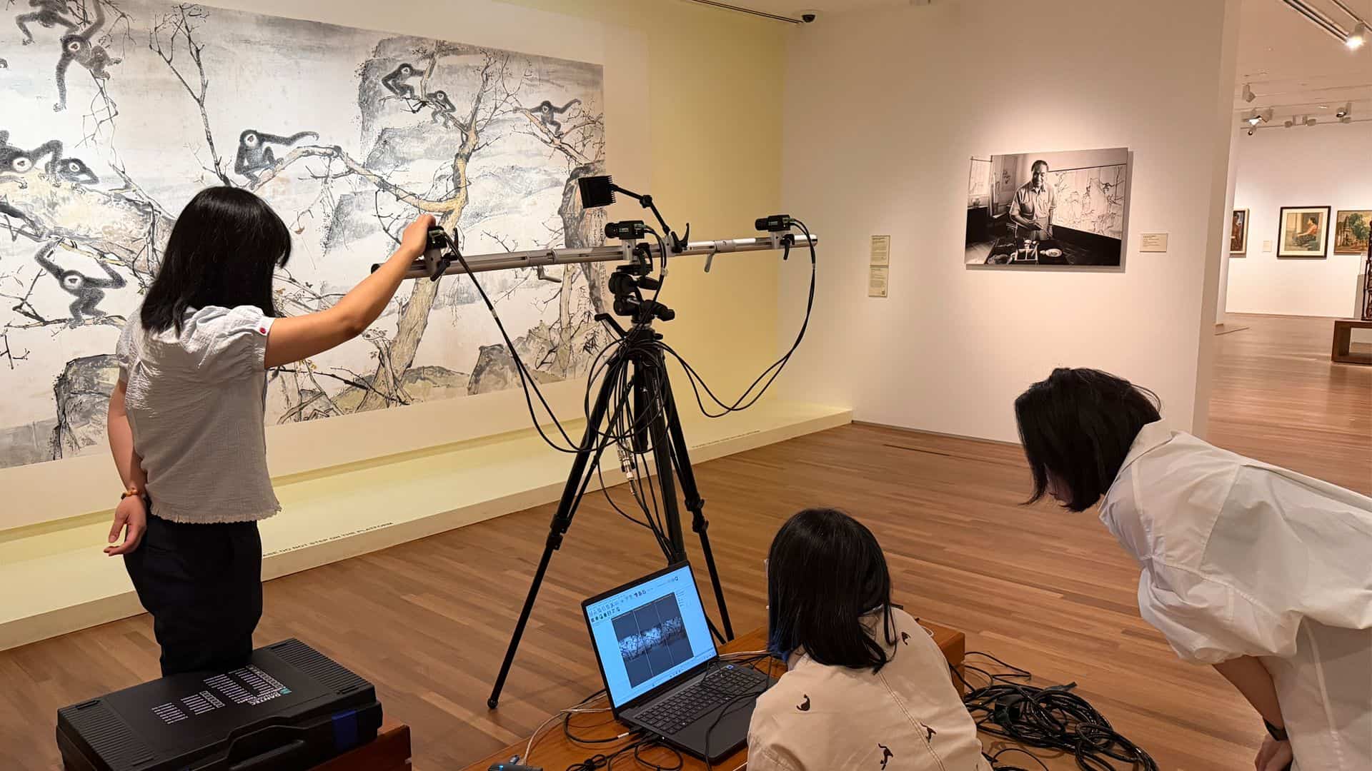 Three people in a gallery use a tripod-mounted camera system to scan an artwork, viewed on a connected laptop screen.