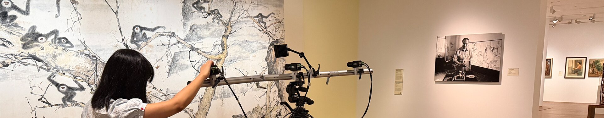 Three people in a gallery use a tripod-mounted camera system to scan an artwork, viewed on a connected laptop screen.