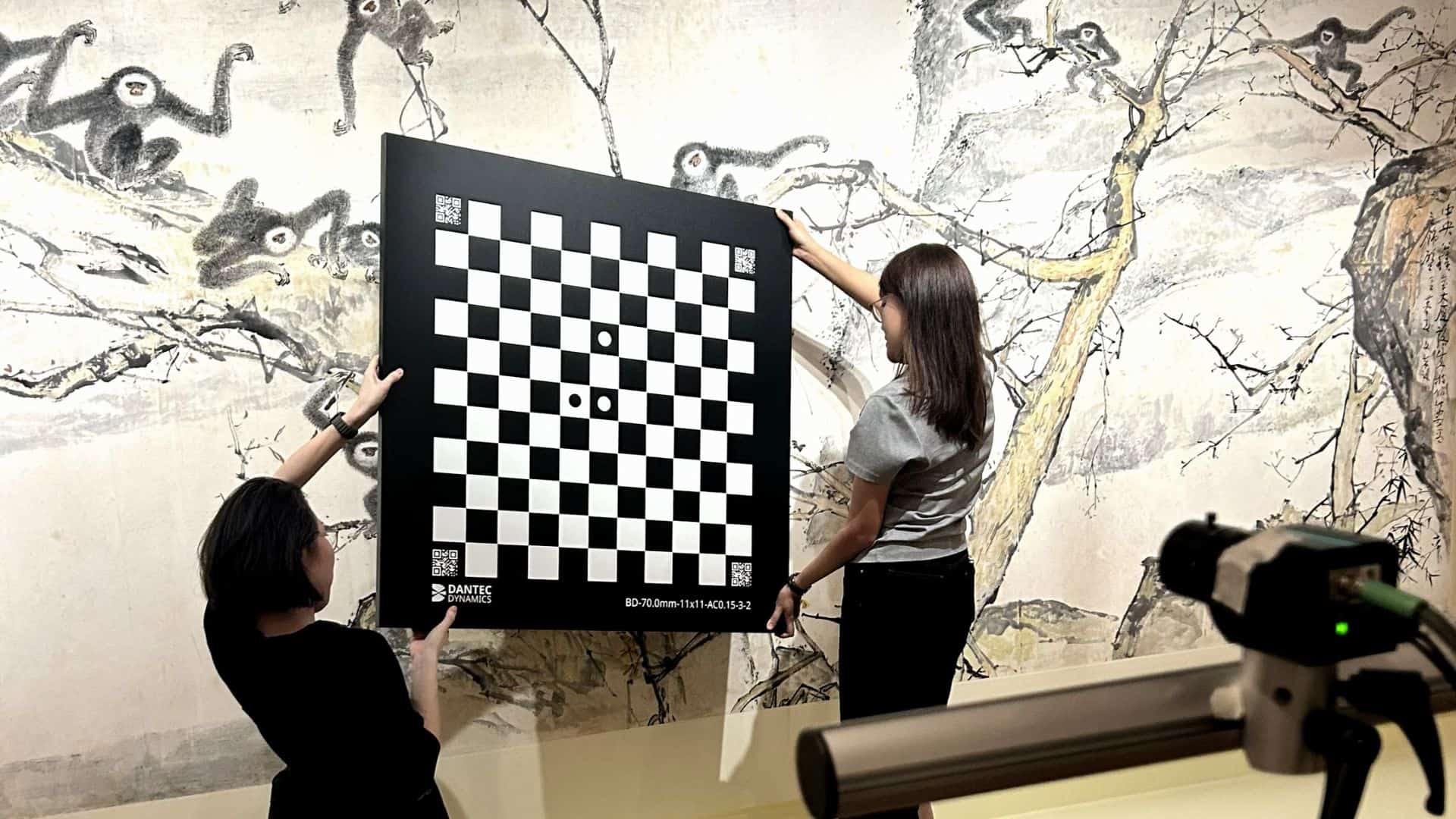 Two women hold a large black and white checkerboard calibration target in front of an artwork.