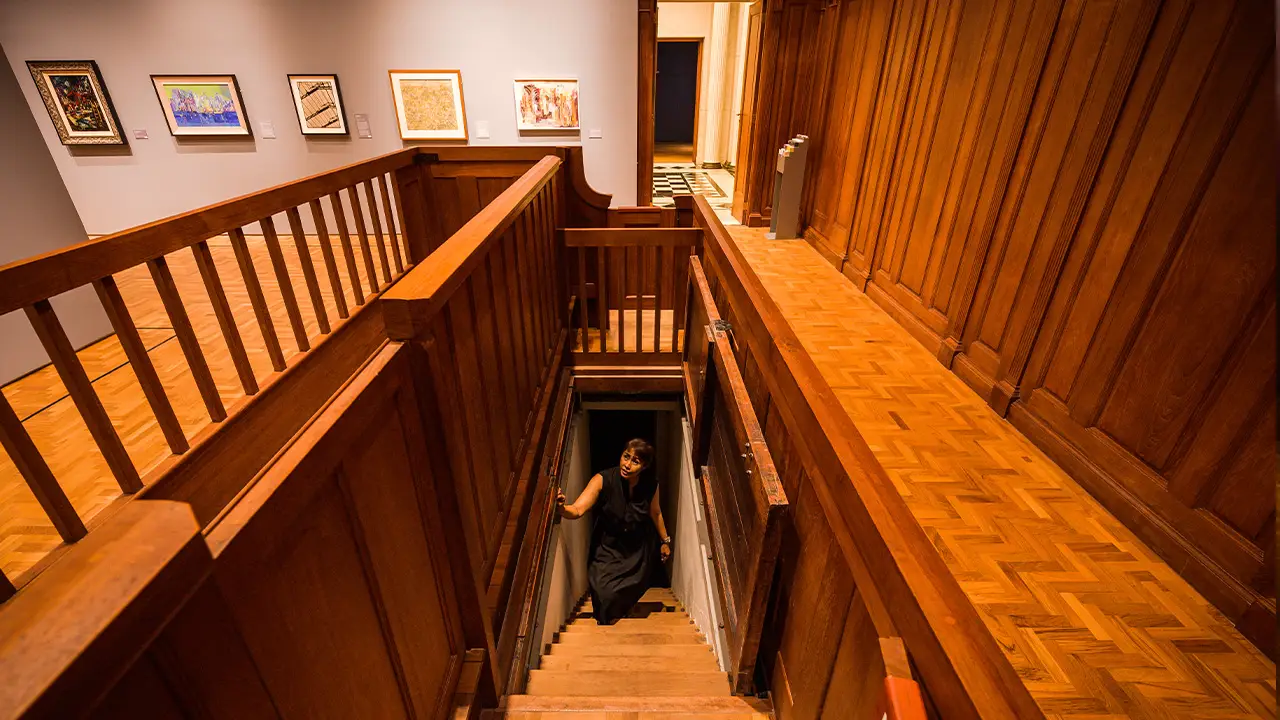 Art gallery interior with wooden staircase, herringbone flooring, and framed artworks on neutral walls as a visitor ascends from lower level