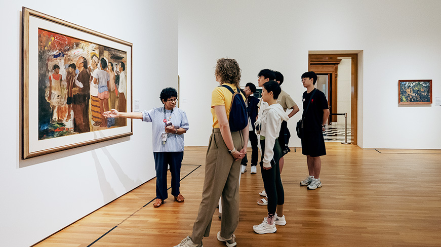 A docent stands in front of a painting explaining the painting's story to a small group of visitors