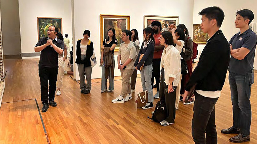 A curator leads a tour in a gallery, speaking to a group of visitors standing in front of framed artworks on the wall