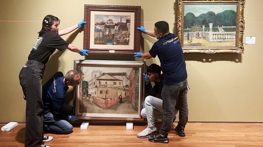 Four art handlers wearing gloves carefully replace a framed painting on the wall