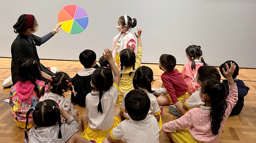 An educator holds up a colour wheel, asking questions to a small group of seated children