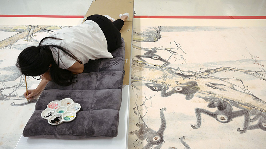 A conservator lies on a padded mat as she delicately restores a large Chinese ink painting of monkeys on tree branches, with a paint palette placed nearby