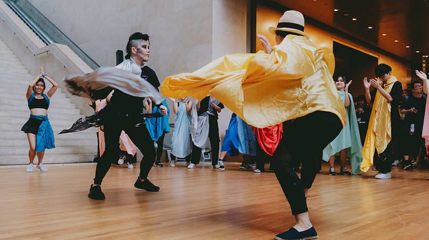 A group of performers in vibrant costumes dance energetically in the Gallery’s atrium, with an audience watching in the background