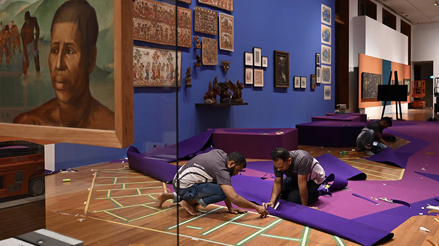 Two exhibition designers placing purple carpeting and taping floor markings for the Tropical exhibition, surrounded by framed artworks