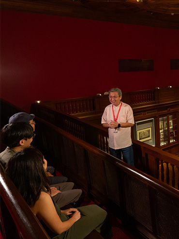 A guide explaining the Back-of-house tour to participants seated at Viewing Gallery benches above the former hearing court