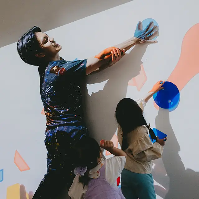A father wearing a shirt splattered with paint and his two daughters having fun in an interactive family art activity 