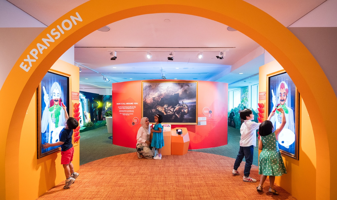 Children play at interactive screens beneath an orange arch in the Keppel Centre for Art Education, with a painting displayed beyond.