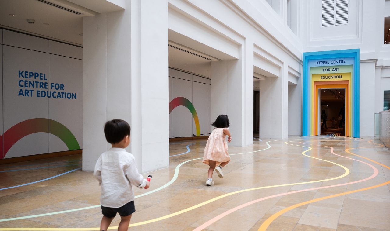 Two young children run along colourful lines on the floor towards the rainbow-framed entrance of the Keppel Centre for Art Education.