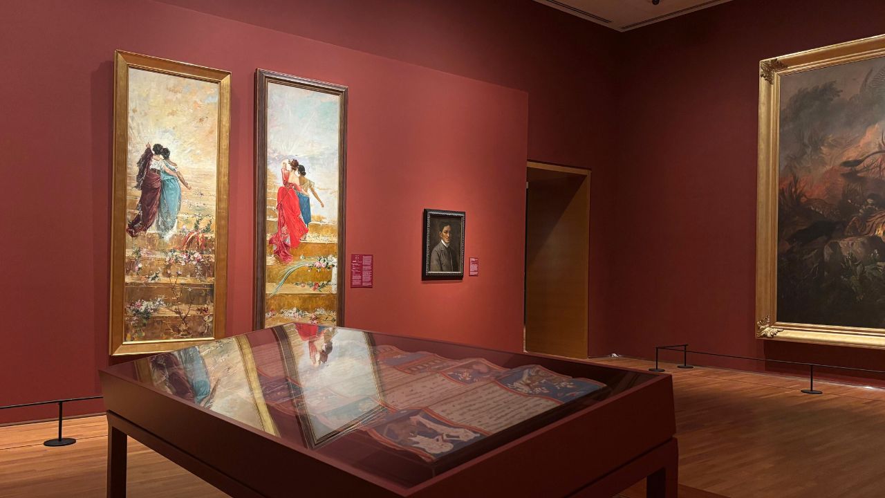 Art gallery room with red walls displaying two large paintings depicting figures in colorful attire. A portrait hangs between them. A display case with documents reflects the artwork, adding depth. Warm lighting enhances a contemplative atmosphere.