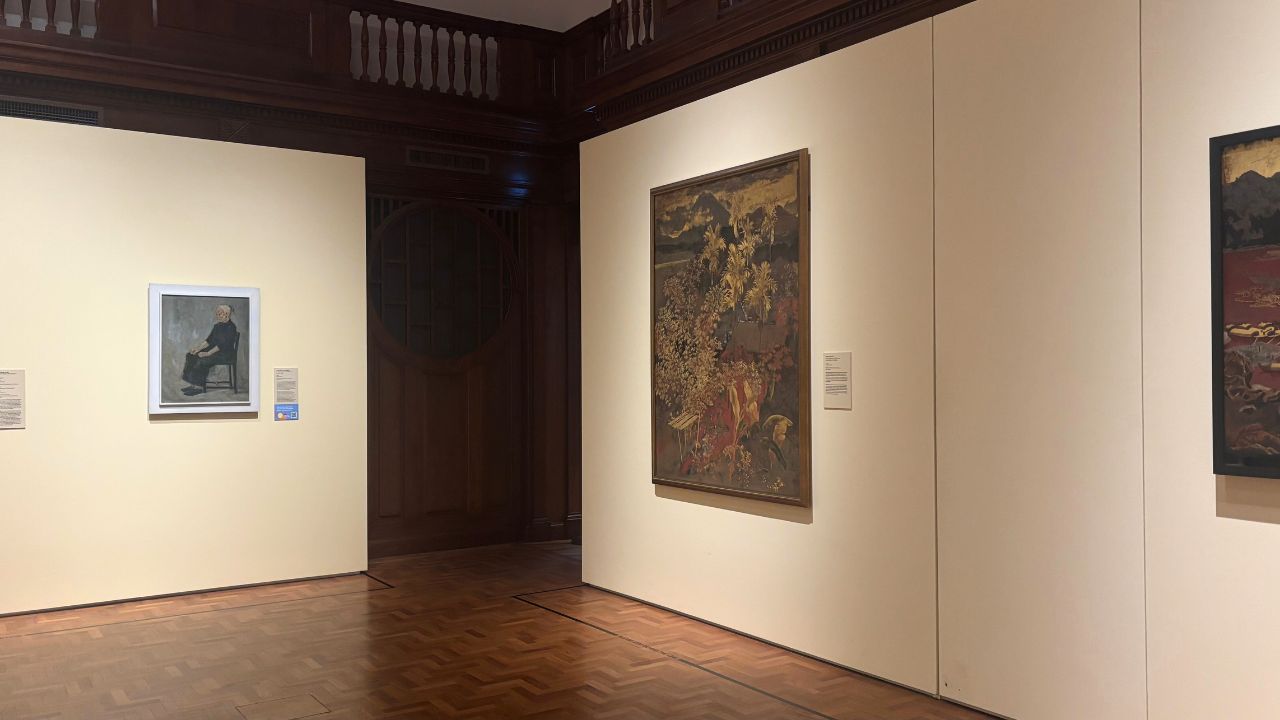 Art gallery room with wooden floors and dark wood paneling. Two large paintings hang on cream-colored walls, each with an informative plaque beside them.