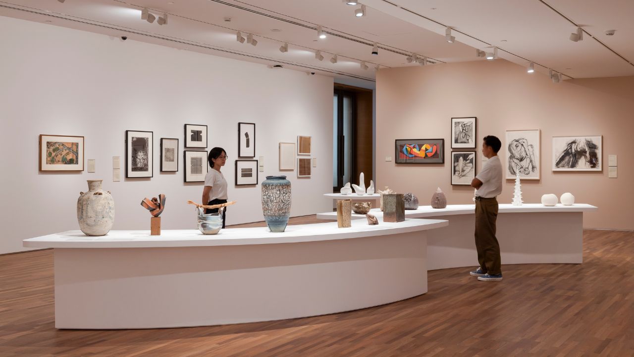 Two visitors view ceramics and framed artworks displayed in a modern art gallery with wooden floors and white walls.