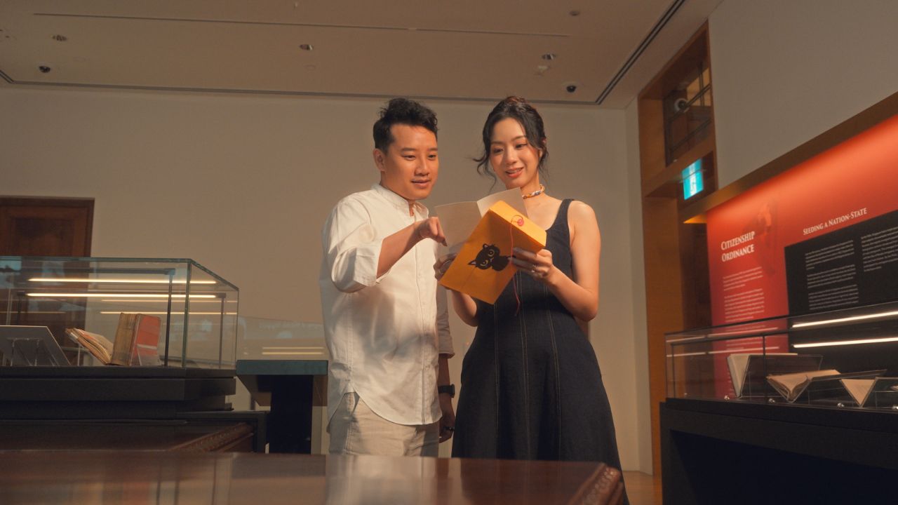 A man and woman are opening an envelope with a puzzle clue, smiling in front of a display case filled with various items.