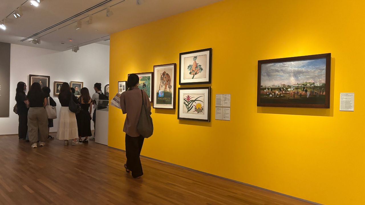 Gallery visitors view framed artwork on vibrant yellow and neutral walls. The atmosphere is contemplative, with people gathered for a closer look.