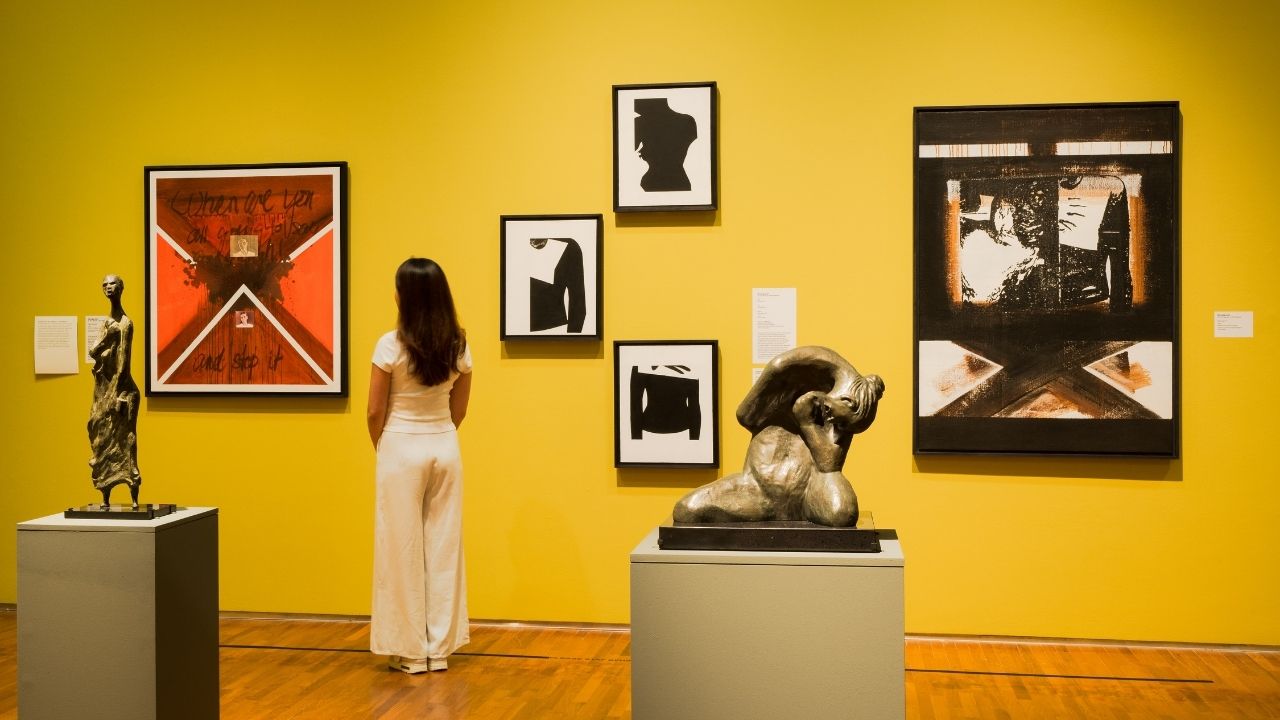 Installation view of Fear No Power, with person standing among paintings and sculptures set against yellow background