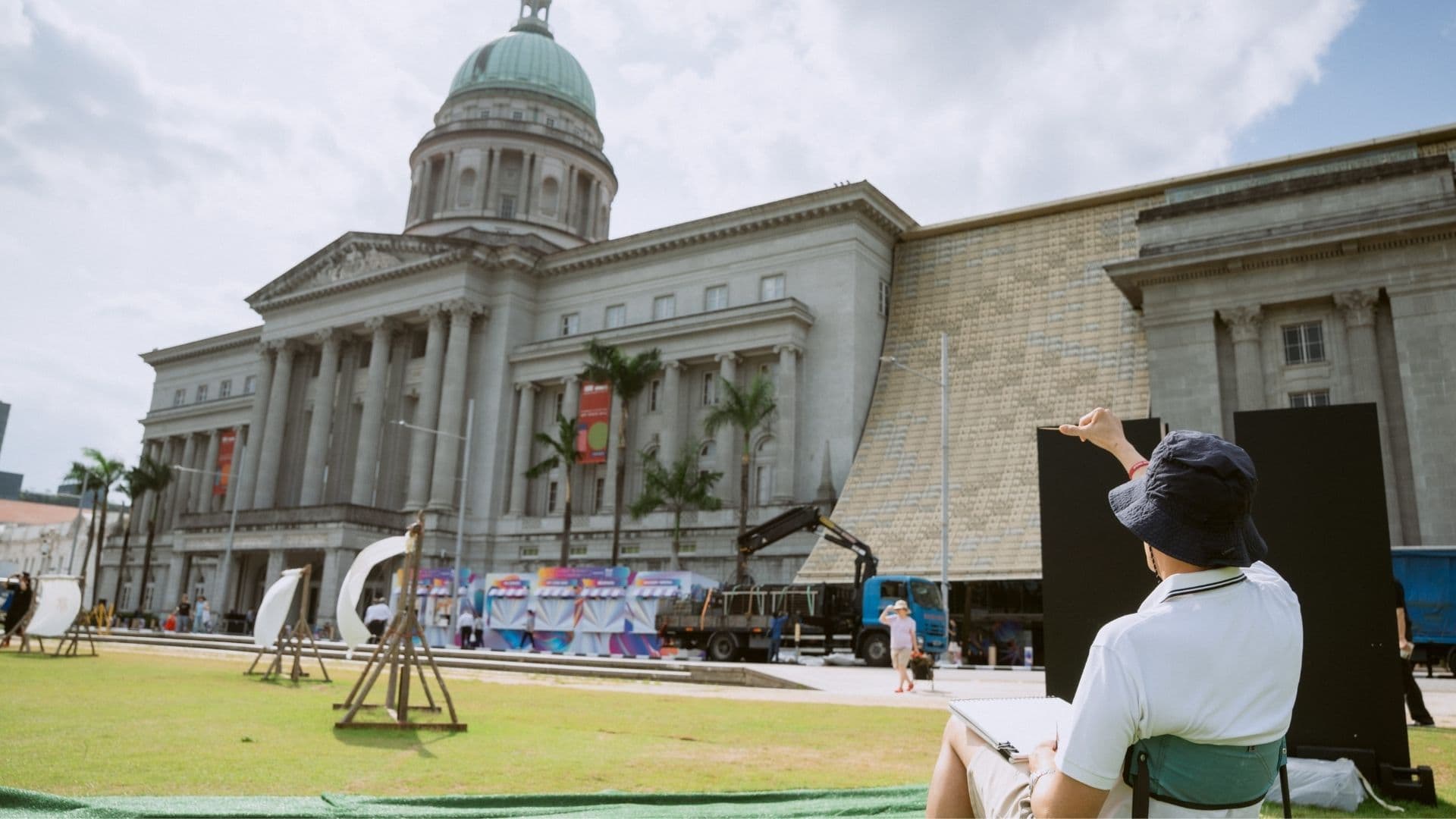 A man sits on a chair sketching in front of a large building, surrounded by trees and a clear blue sky.