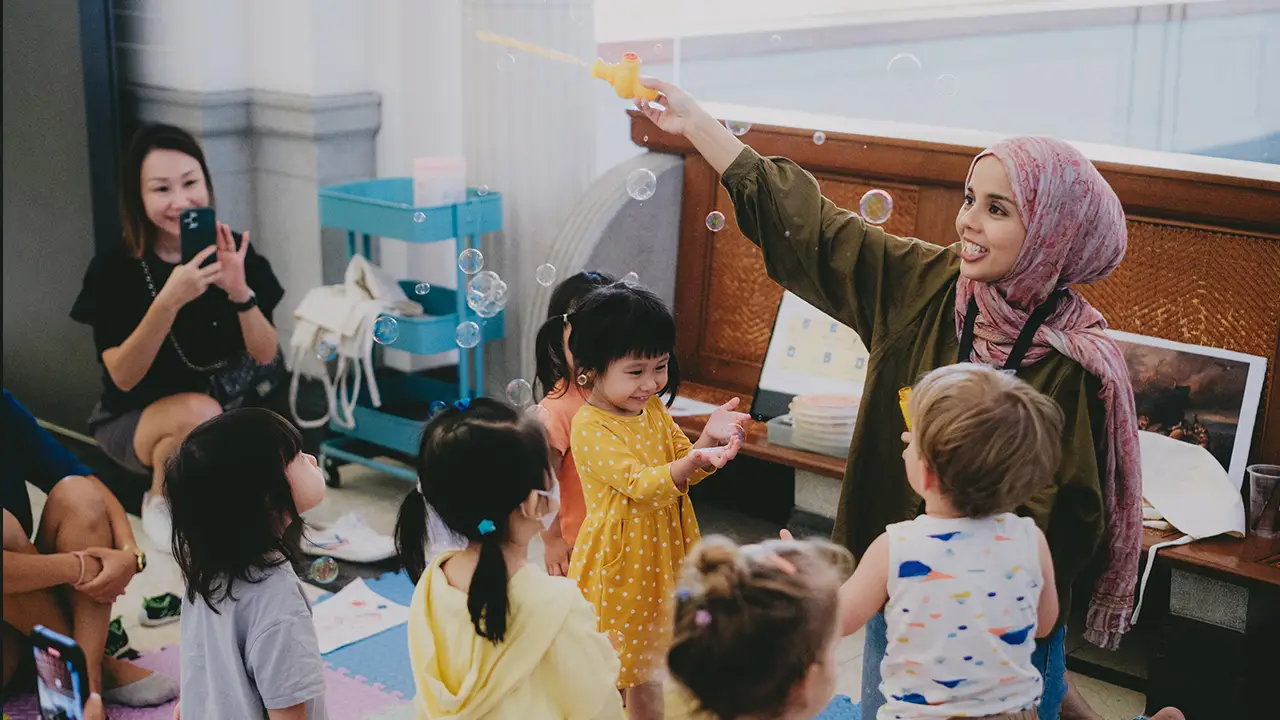 GalleryToddlers!-Facilitator blowing bubbles while toddlers gather around during a playful activity session in the gallery.