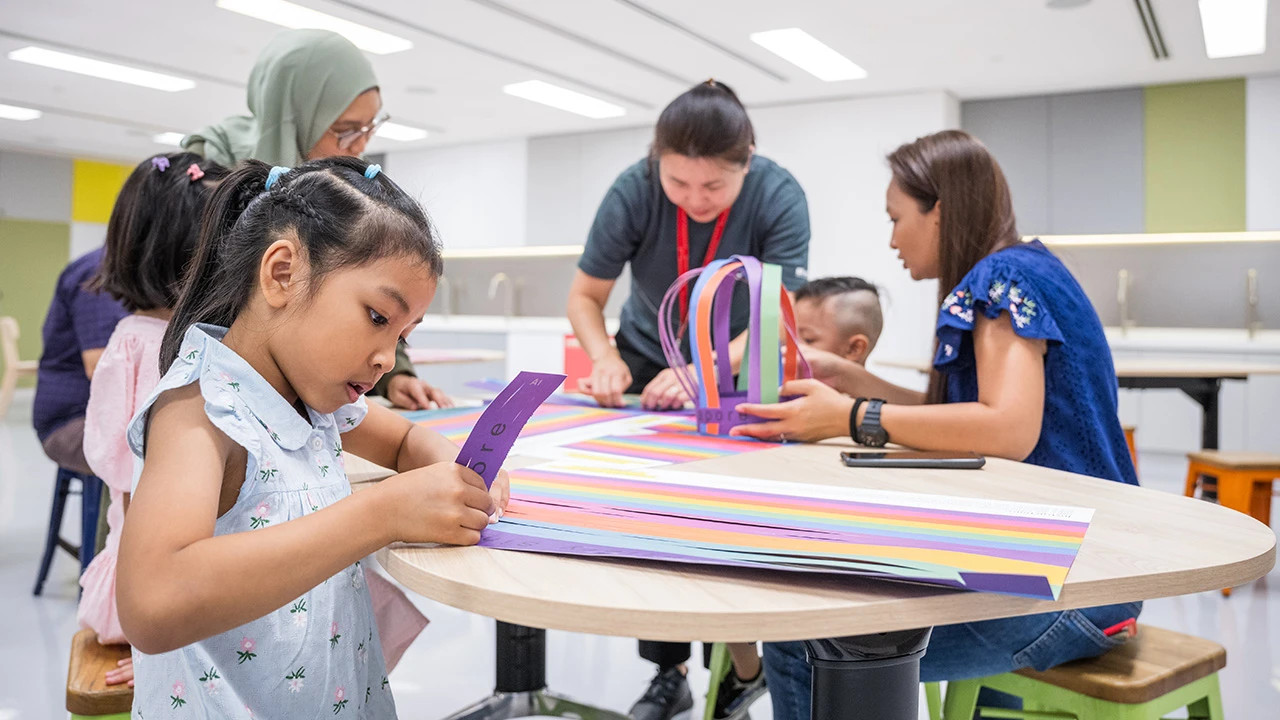 Children crafting with strips of colourful paper, alongisde staff and guides from the Keppel Centre for Art Education. The room they are seated in is bright, with grey walls and neutral lighting. 