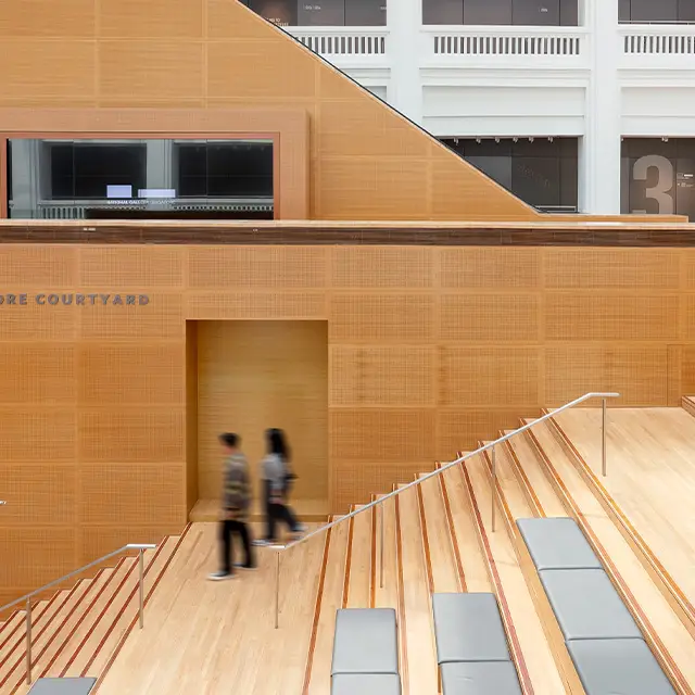 A man and woman walk down a grand wooden staircase in a spacious gallery with an escalator in the background