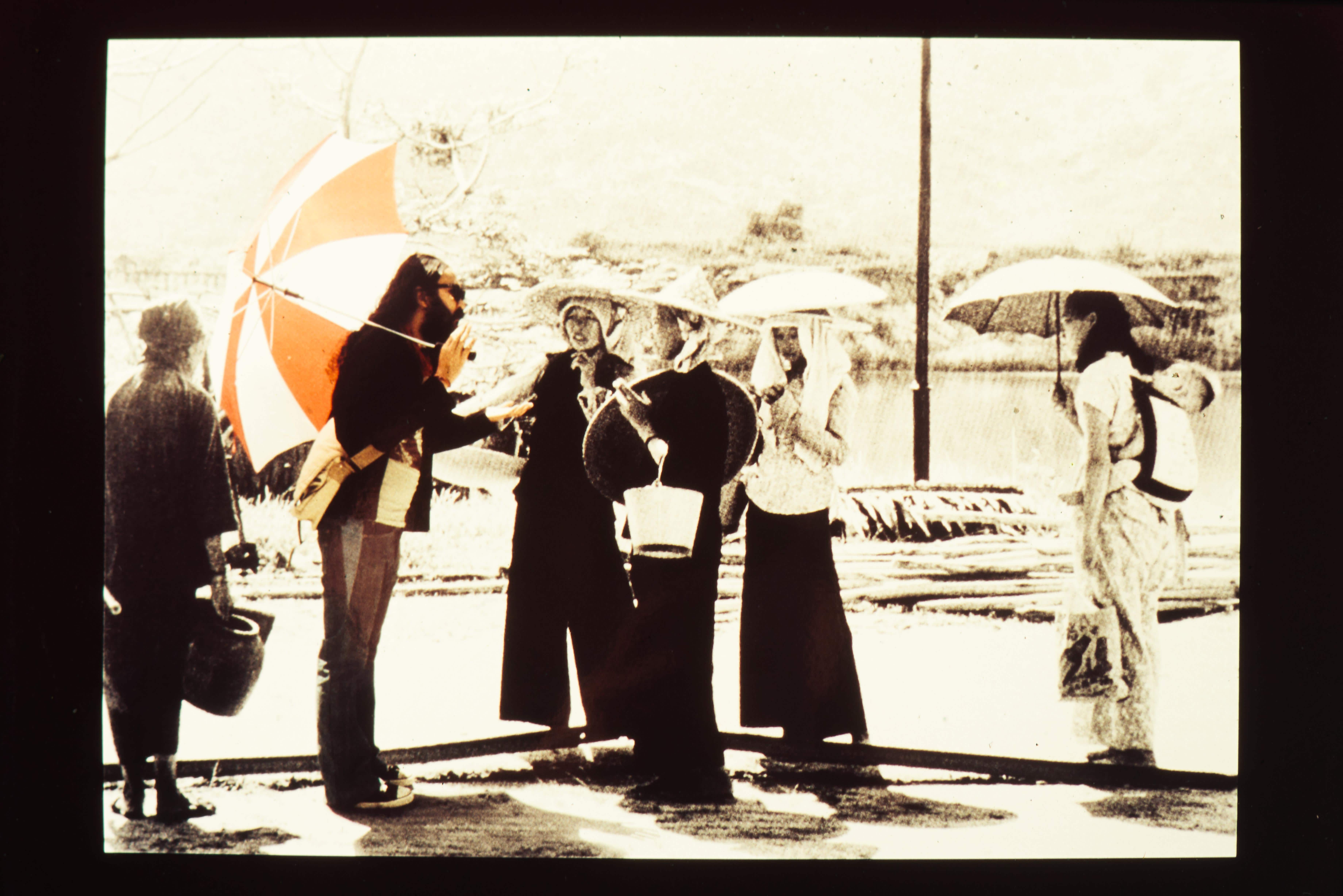 Women in black and white, wearing caps and holding an umbrella, stand next to a man depicted in color.