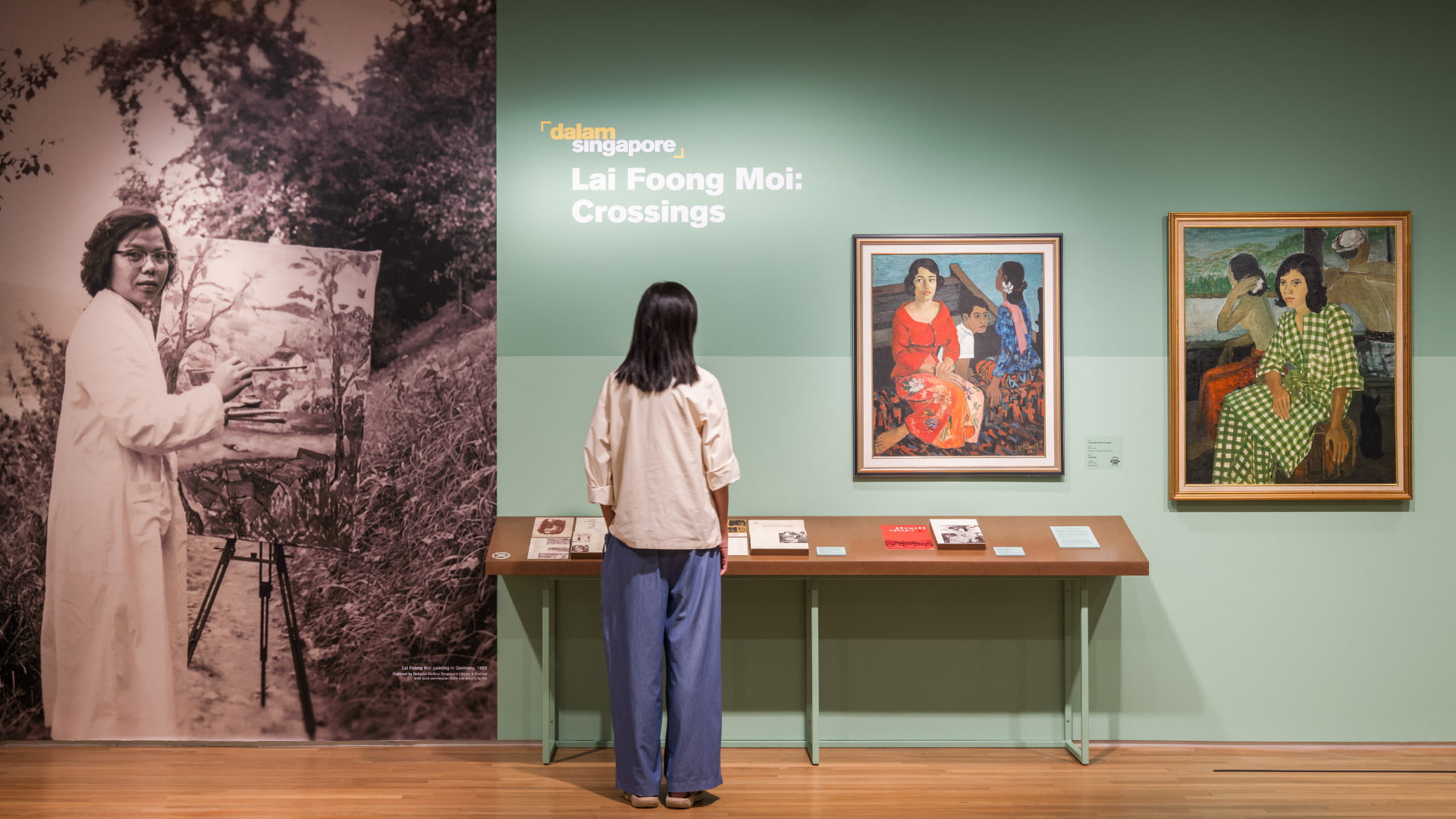 Promotional graphic for the exhibition Lai Foong Moi: Crossings featuring a painting by Singapore artist Lai Foong Moi. The artwork depicts three figures seated together, with a woman in a red blouse and floral-patterned skirt in the foreground, alongside two younger figures dressed in blue and white. The background uses deep teal and earthy tones. Exhibition title text appears on the right against a muted green background with the Dalam Singapore logo above.