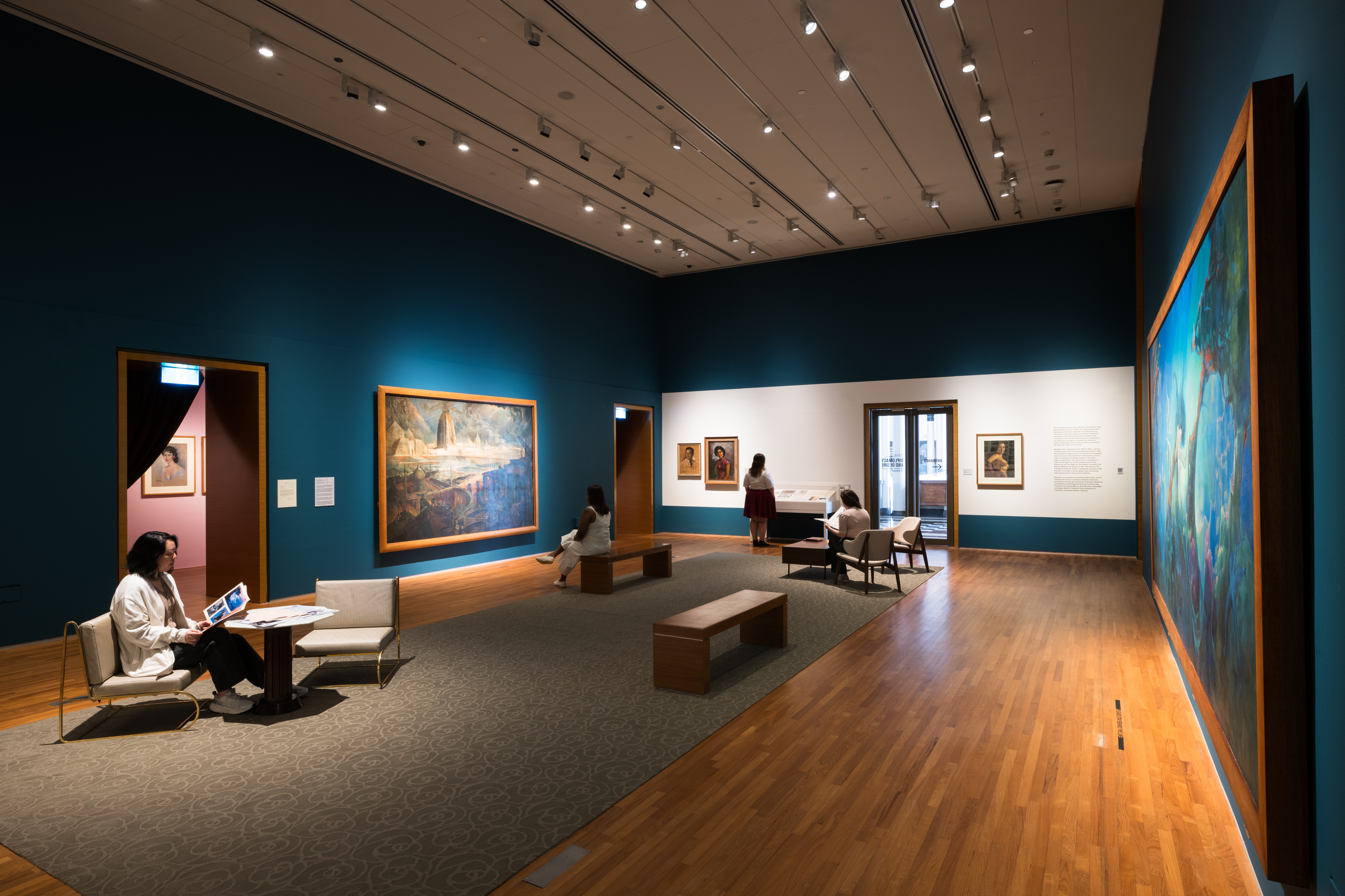 Visitors seated and walking through a contemporary art gallery with teal walls, wooden flooring, and framed paintings on display