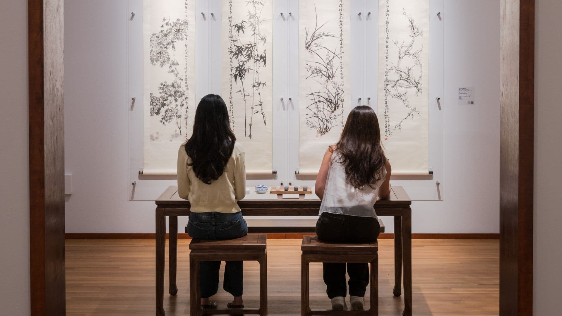 Two women seated at a table, engaged in discussion while observing paintings by artist Fan Chang Tien on the wall.