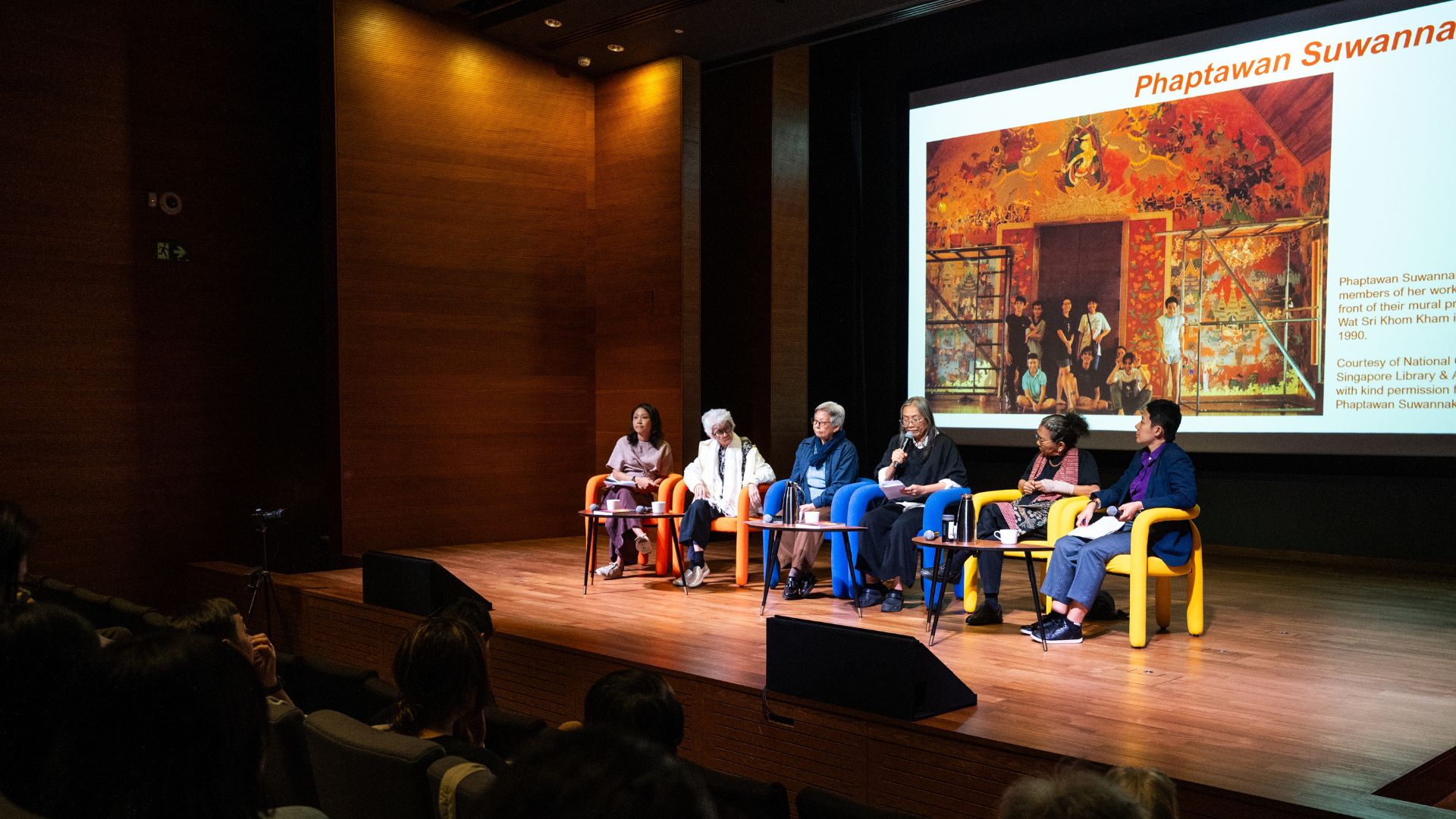 A panel of six people sits on a stage with vibrant colored chairs, engaged in a discussion. A large screen displays artwork and the name "Phaptawan Suwannakudt." The atmosphere is focused and attentive.