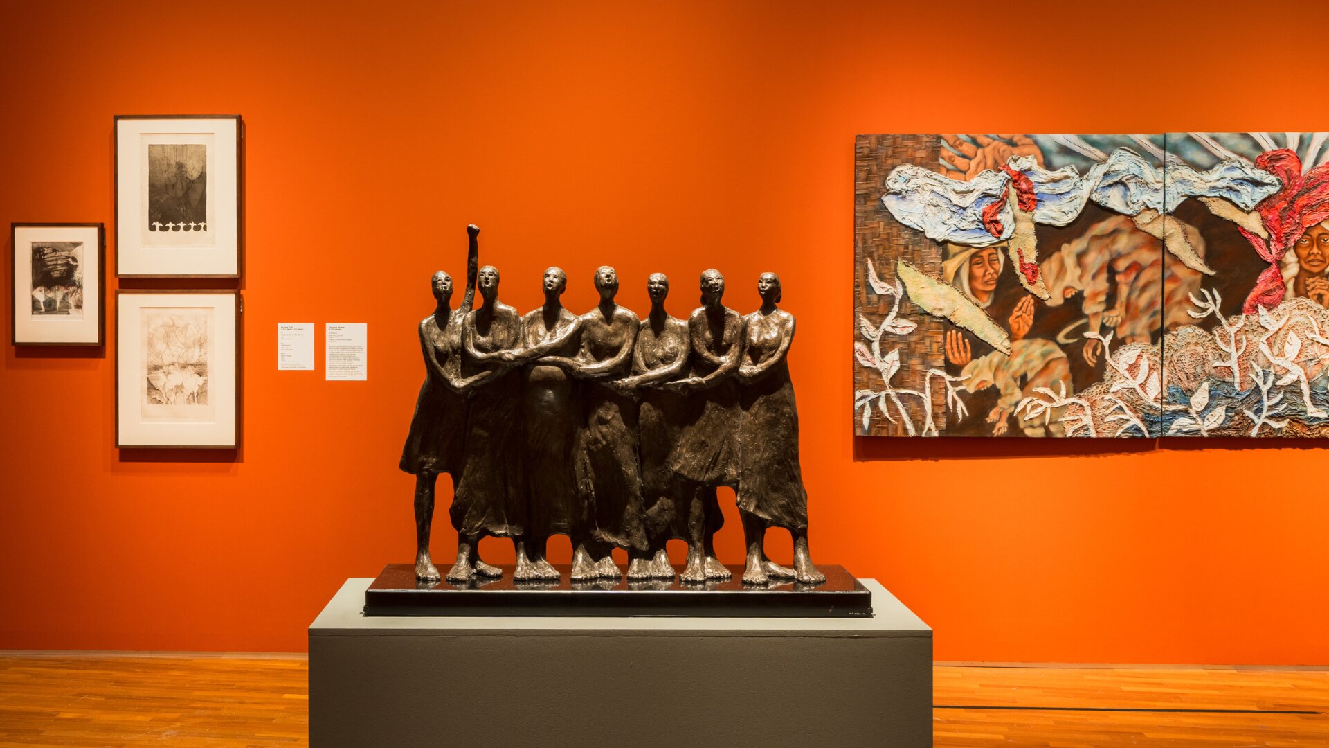 Sculpture depicting women holding hands in unity, displayed in a museum alongside vibrant paintings.