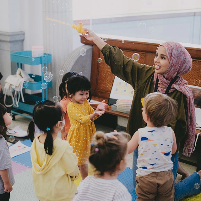 A smiling woman in a headscarf playfully blows bubbles for a group of happy young children gathered around her