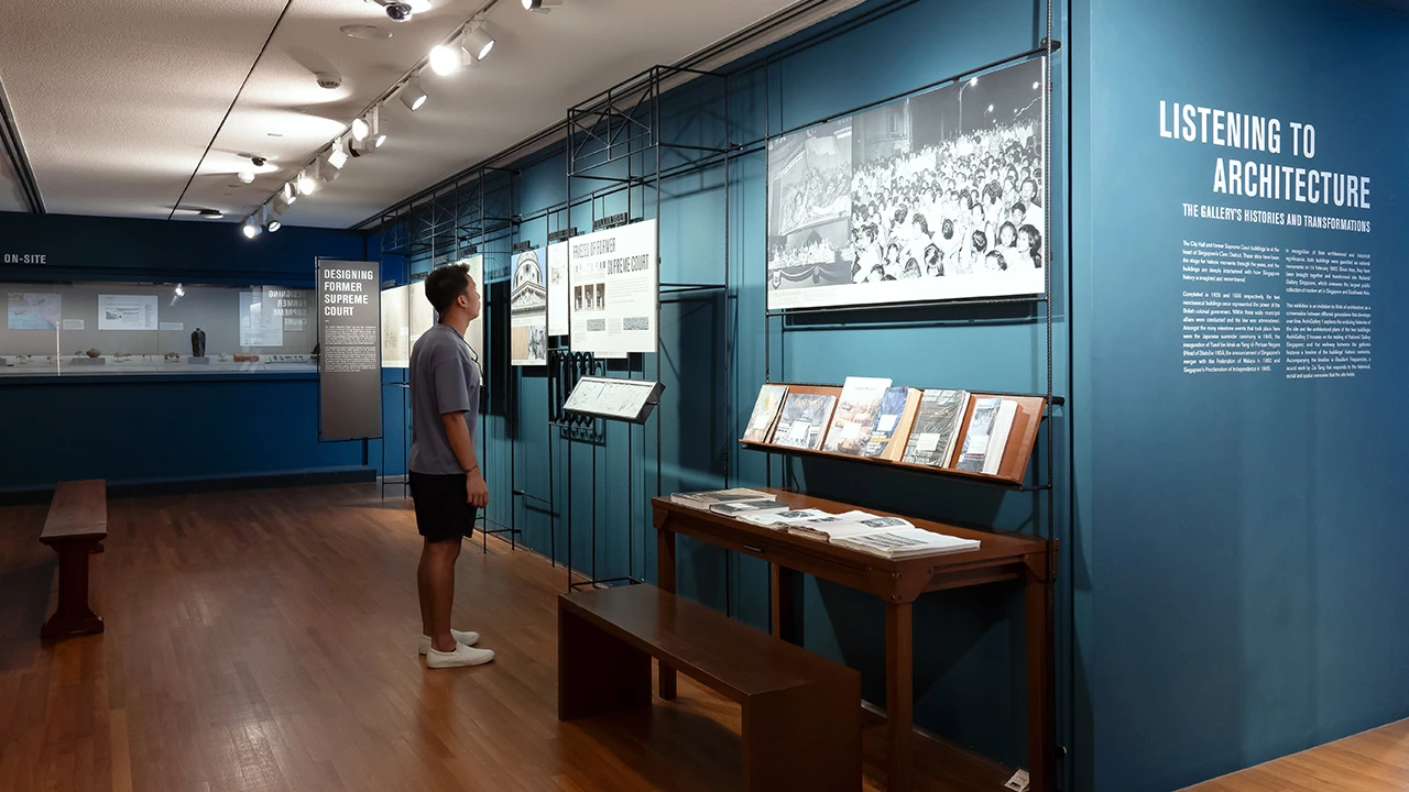 A visitor stands reading interpretive displays in a museum gallery with blue walls, featuring photographs, books and exhibition panels about architecture. Wooden benches and display tables line the space beneath track lighting, creating a quiet environment for exploring the exhibition.