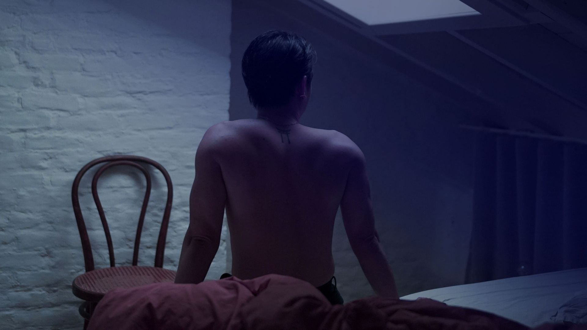 A man is seen in a shadowy bedroom, standing next to a chair, evoking a sense of intrigue.