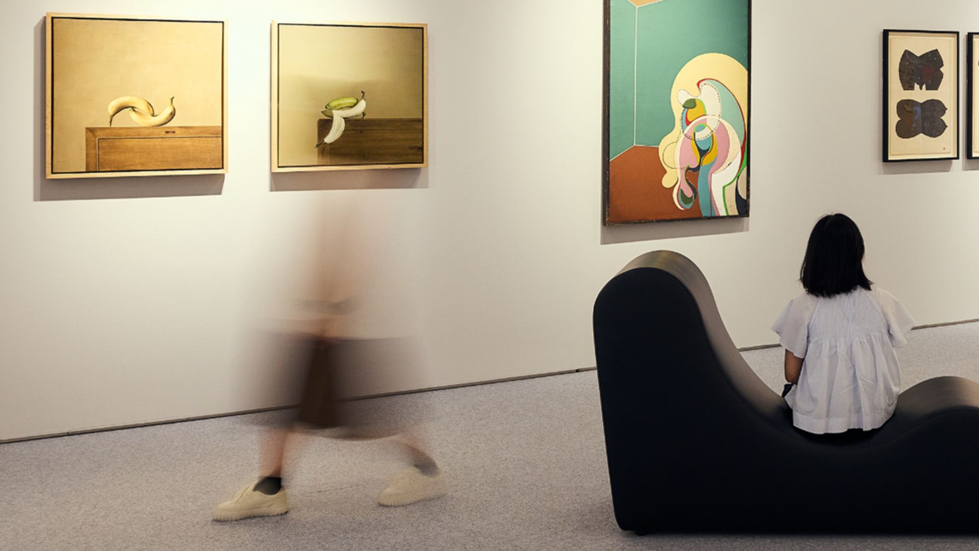 A woman seated on a chair in an art gallery, surrounded by various artworks displayed on the walls.