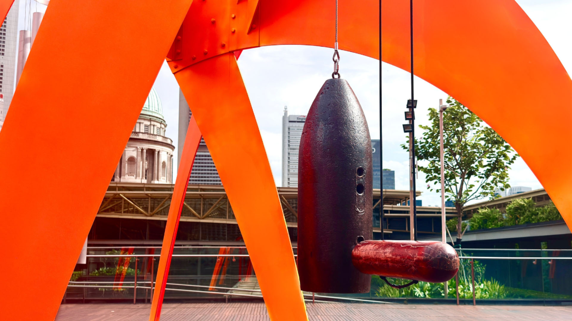 A large orange sculpture made from alloys of defused unexploded ordnance, with windchimes suspended from it, creating a striking visual contrast
