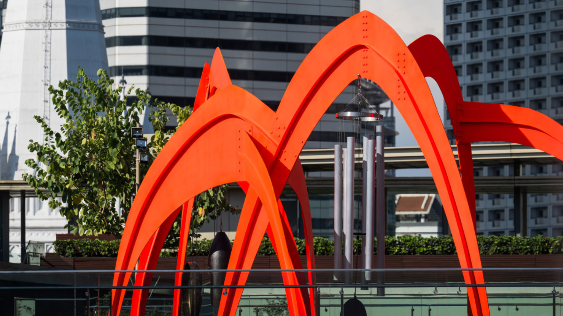 Large orange-painted art installation in the shape of arches, with large wind chimes hanging from its frame.