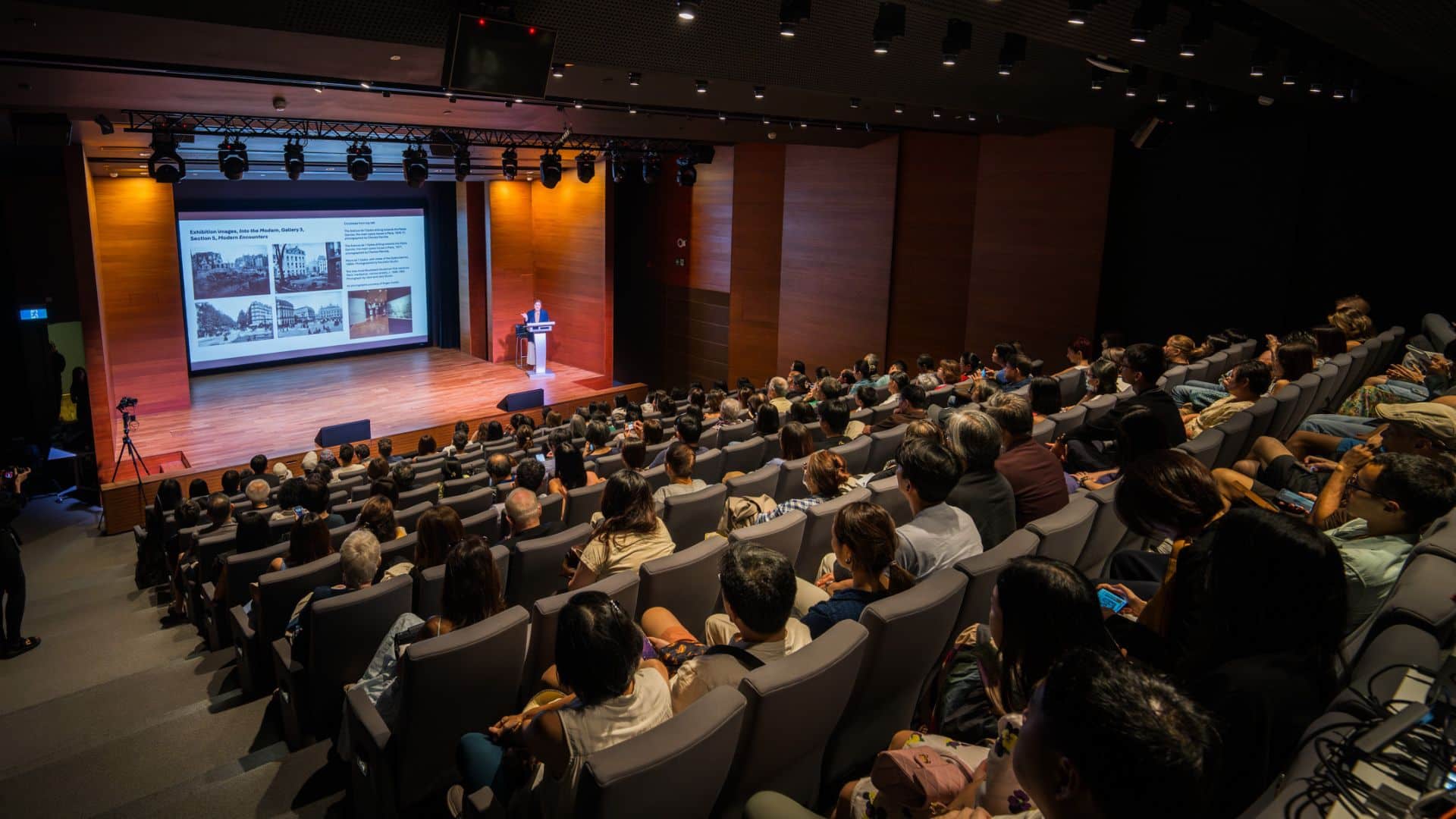 A large room filled with seated attendees as a man presents at the front, engaging the audience with his talk.