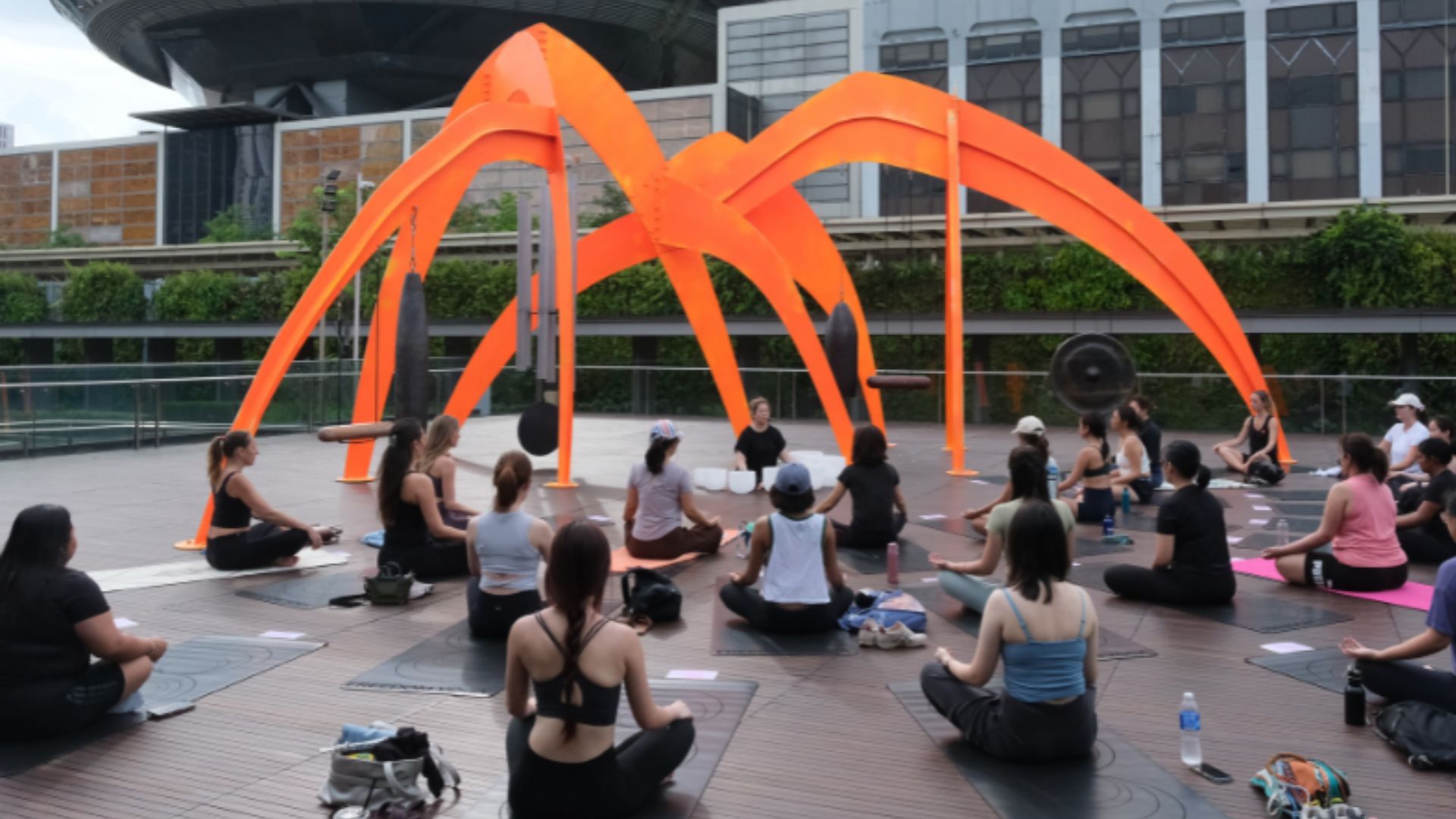 Several individuals meditating on the ground, facing an abstract orange sculpture, creating a vibrant outdoor scene.