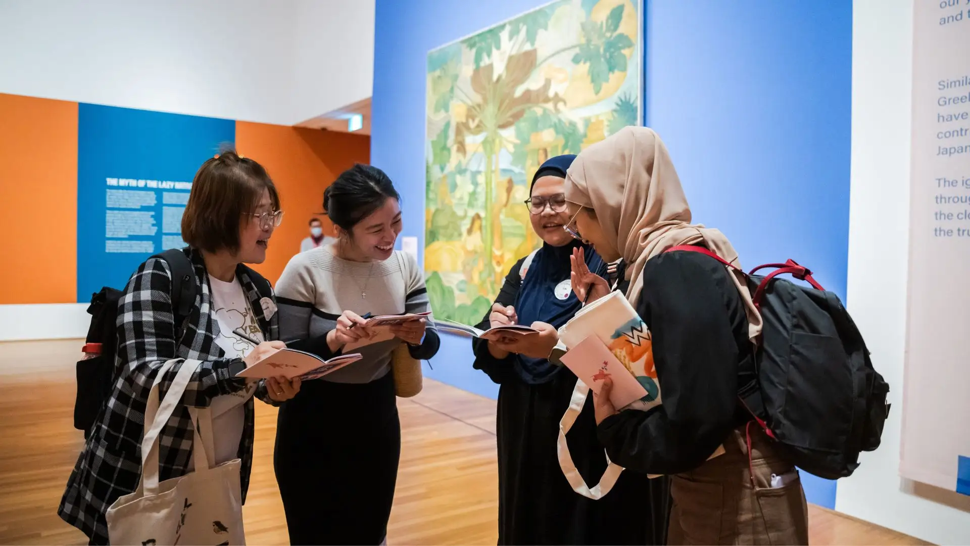 Four friends standing inside an art gallery and laughing together. They are holding activity booklets and talking about the activity.