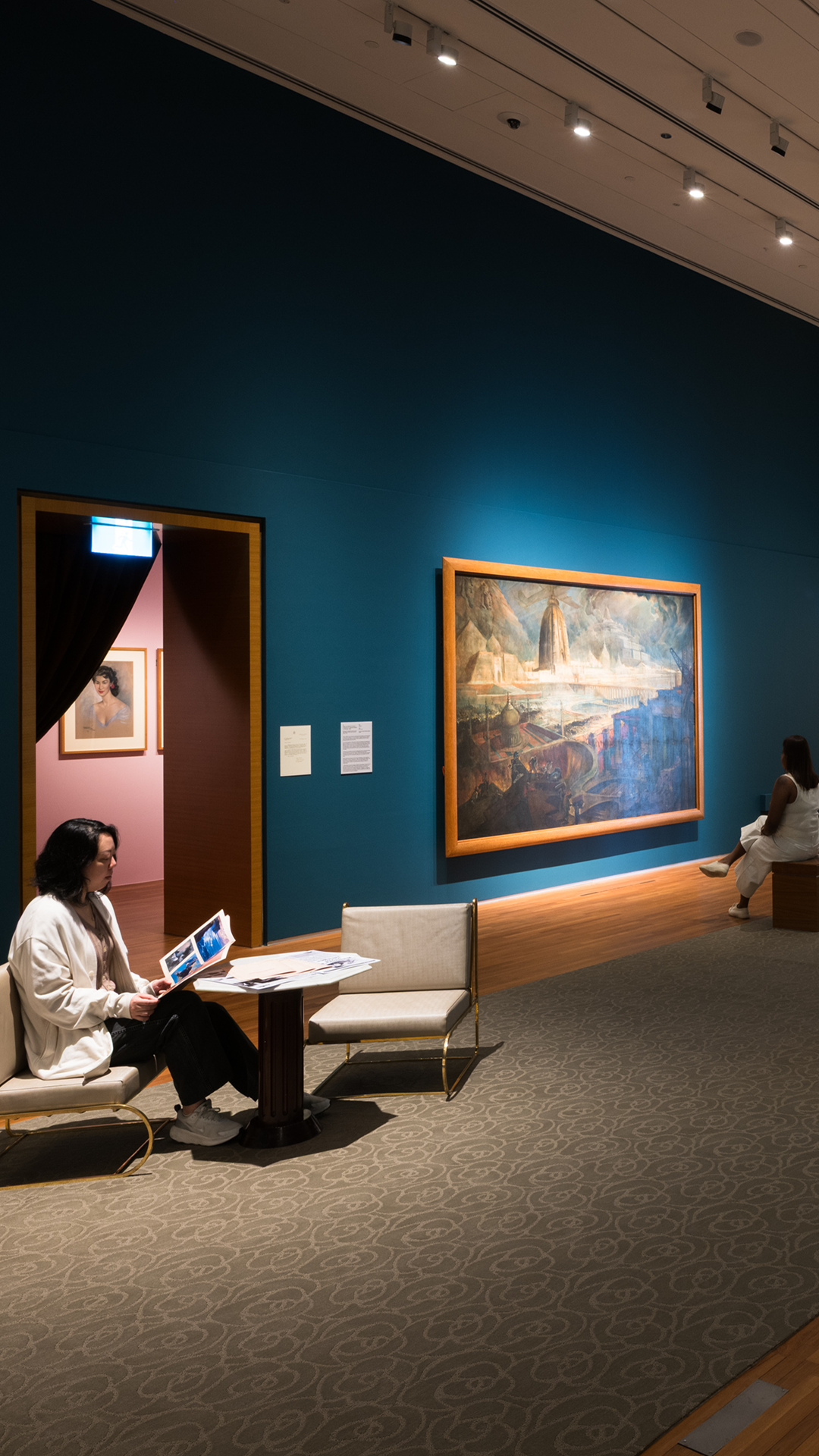 Seated visitor in a teal-walled gallery and another visitor viewing a large landscape painting of mountains and waterfall in a calm exhibition setting