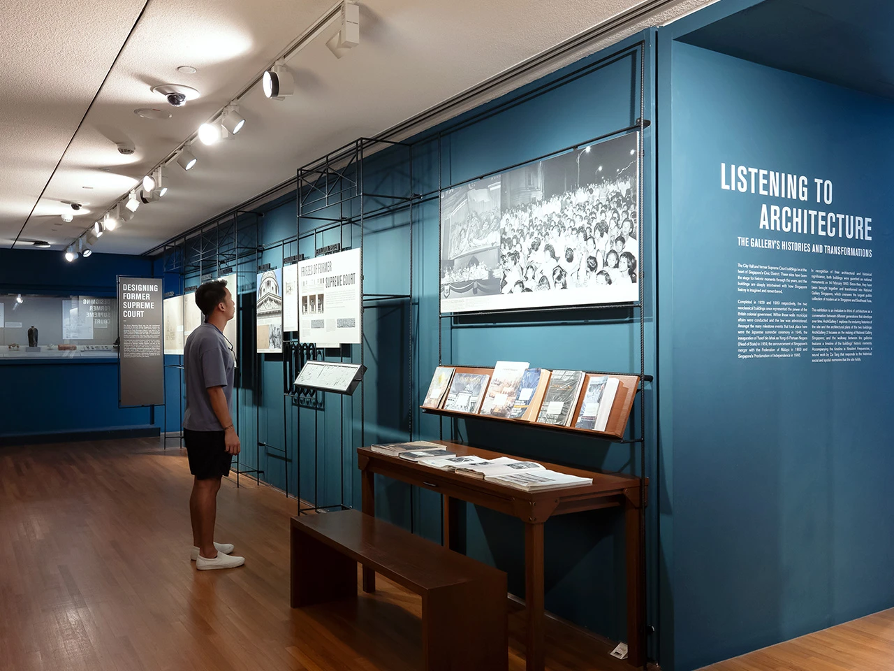 Visitor observing “Listening to Architecture” exhibition exploring sound and history in a blue-walled gallery setting