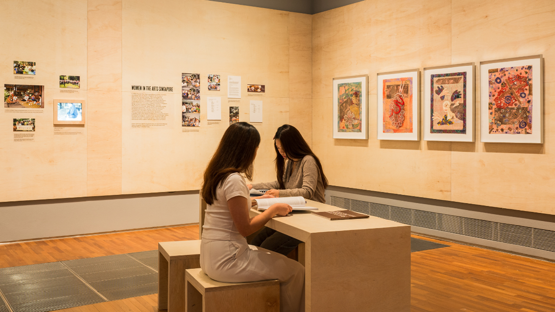 Two women seated at a table in an art museum, reading exhibition texts amidst a backdrop of artwork.