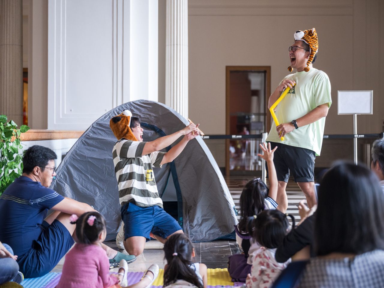 Families gather for an interactive storytelling session in a museum atrium. Two performers wearing tiger hats act out a scene beside a small camping tent, while children sit on the floor with raised hands and adults watch from the audience.