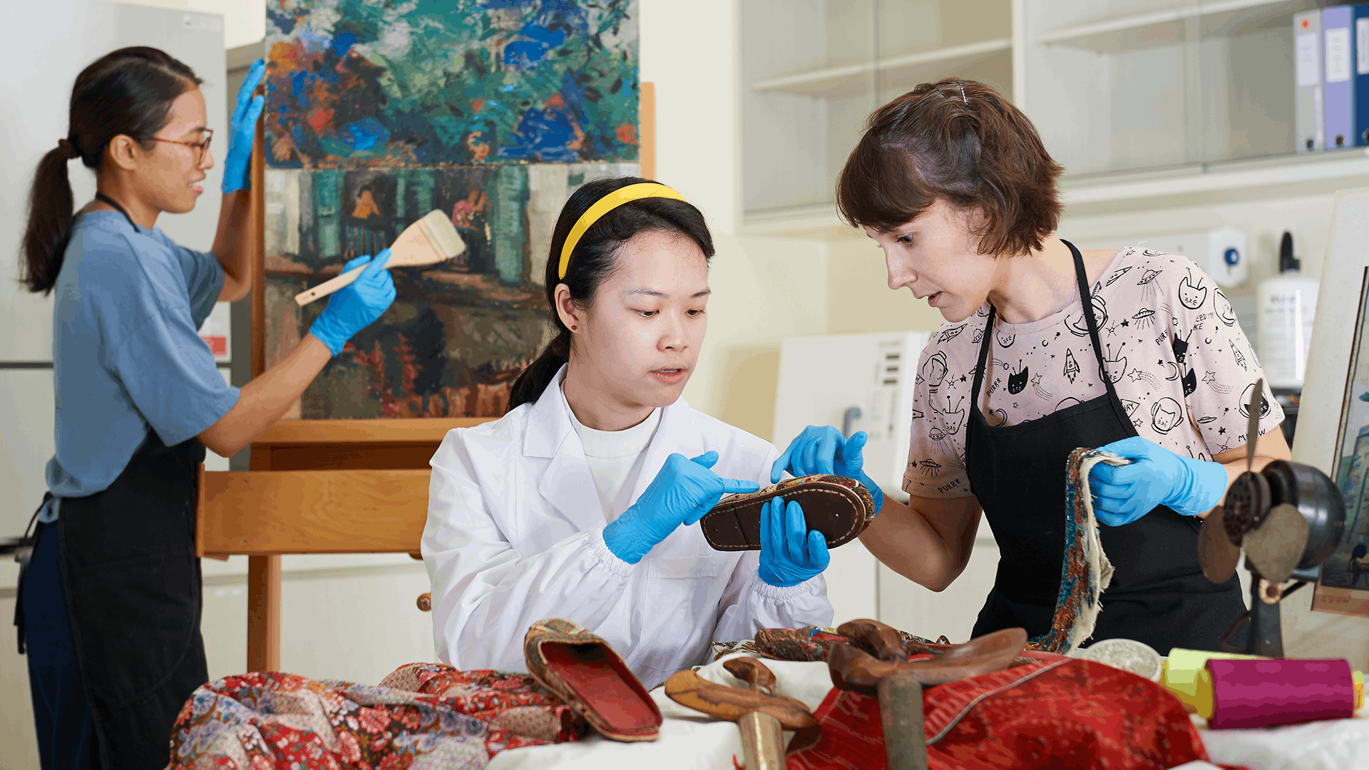 Three art conservators examining paintings, artefacts and archival material.
