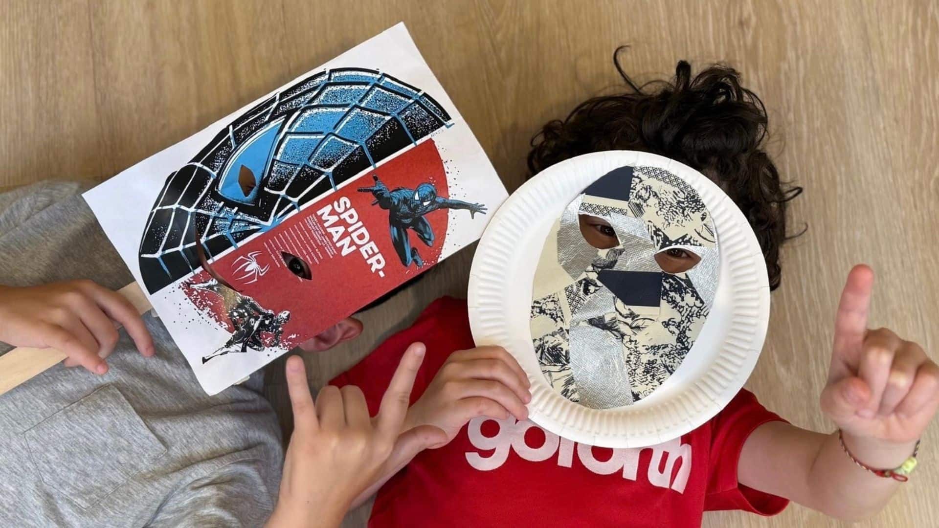 Two kids hold up paper plates decorated with Spider-Man masks, showcasing their creative art project.