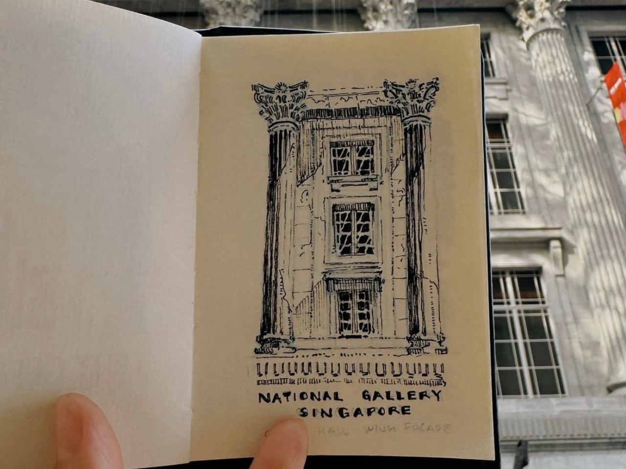Close-up of a sketchbook page featuring a black ink illustration of a classical building façade with columns and windows, above the words “National Gallery Singapore.” A finger is visible holding the page open, with part of a real building exterior blurred in the background.